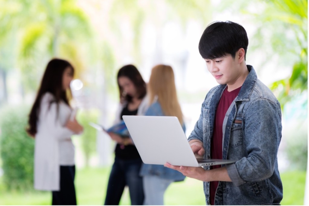 Student using a laptop outdoors while classmates collaborate in the background, representing digital learning and connected education.